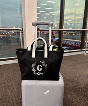 Cargar imagen en el visor de la galería, Black embroidered boat tote bag with white handles and a white letter G on front, placed on a gray suitcase