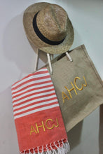 Load image into Gallery viewer, Personalized beach towel with red stripes and initials AHCJ hanging next to a straw hat and beige tote bag