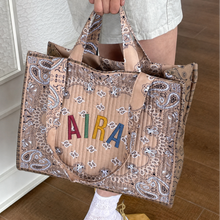 Load image into Gallery viewer, A person holds a large tan quilted tote bag with a paisley print and colorful embroidered letters spelling 'AIRA' on the front, shown in an indoor setting with wood flooring.
