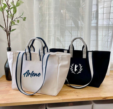 Two embroidered boat tote bags by Travellers Hat Boutique with personalized designs on a wooden table