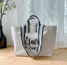 Lataa kuva Galleria-katseluun, White embroidered boat tote bag with navy blue striped handles on wooden surface
