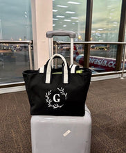 Lataa kuva Galleria-katseluun, Black embroidered boat tote bag with white handles and a white letter G on front, placed on a gray suitcase
