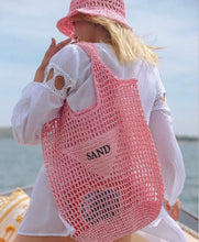 Carregar imagem no visualizador da galeria, Handmade raffia bucket bag in pink mesh style with SAND patch carried by woman in white dress at the beach
