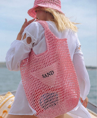 Handmade raffia bucket bag in pink mesh style with SAND patch carried by woman in white dress at the beach