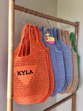 Carregar imagem no visualizador da galeria, Handmade raffia bucket bag in orange, blue, beige, and green hanging on a wooden rack by Travellers Hat Boutique

