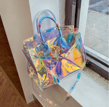 Carregar imagem no visualizador da galeria, Holographic clear tote bag with blue handles from Travellers Hat Boutique displayed on a windowsill
