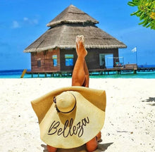 Lade das Bild in den Galerie-Viewer, Person relaxing on beach wearing oversized straw sun hat with Belleza text near tropical water bungalow
