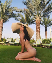 Lade das Bild in den Galerie-Viewer, Woman wearing oversized straw sun hat kneeling on grass with palm trees and lounge chairs in background
