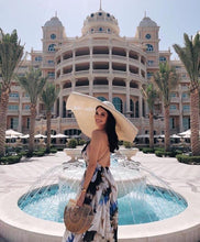 Lade das Bild in den Galerie-Viewer, Woman wearing oversized straw sun hat and floral dress standing by fountain in front of elegant building
