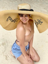 Lade das Bild in den Galerie-Viewer, Woman wearing oversized straw sun hat with wide brim and black ribbon sitting on sandy beach
