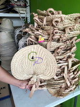 Load image into Gallery viewer, Hand holding a personalized embroidered abaniko fan with ribbons in front of stacked woven fans
