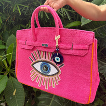 Carregar imagem no visualizador da galeria, Bright pink terry beach bag with eye design and charm held by hand in outdoor greenery
