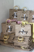 Load image into Gallery viewer, Travellers Hat Boutique personalized tote bags with bamboo handles in beige fabric stacked on shelves
