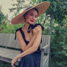 Lade das Bild in den Galerie-Viewer, Woman sitting on a bench wearing a large wheat straw hat with a black ribbon under the chin
