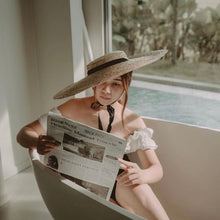 Lade das Bild in den Galerie-Viewer, Woman wearing a wide-brimmed straw hat with black ribbon sitting in a bathtub reading a newspaper near a window
