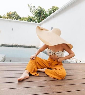 Woman wearing an oversized straw sun hat sitting on wooden deck by a pool in casual summer outfit