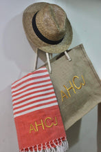 Load image into Gallery viewer, Personalized beach towel with red stripes and initials AHCJ hanging next to a straw hat and beige tote bag
