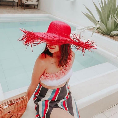 Woman wearing a vibrant red fringe sun hat for women sitting by a pool with a striped skirt and floral top