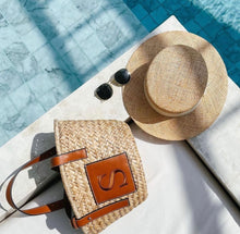 Lade das Bild in den Galerie-Viewer, Seagrass basket handbag with leather handles and monogram near a straw hat and sunglasses by the pool

