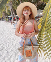 Lade das Bild in den Galerie-Viewer, Woman on beach holding seagrass basket handbag with leather straps and monogram detail
