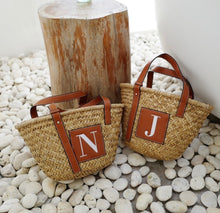 Lade das Bild in den Galerie-Viewer, Seagrass basket handbag tote with brown leather handles and initial patches on white pebble ground
