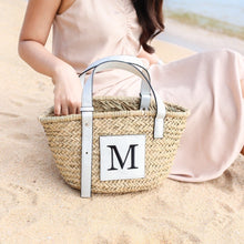 Lade das Bild in den Galerie-Viewer, Seagrass basket handbag with white leather handles and monogram M patch held by woman on sandy beach
